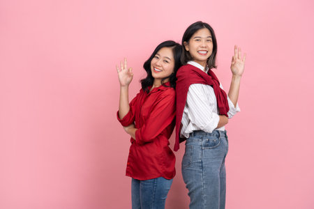 Two happy beautiful Asian girls holding ok sign and looking at camera over pink background. Two people conceptの写真素材