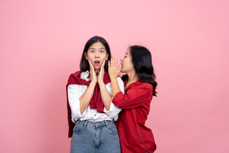 Two excited Asian girls in casual shirts whispering secrets isolated on pink background