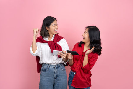 Two cheerful Asian girls friends standing alone on a pink background. They are playing games on their mobile phones.の写真素材