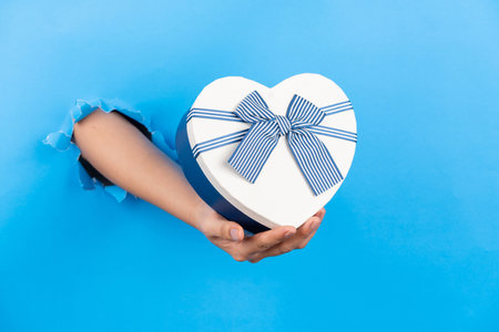 A woman's hand reaches out of a hole on a blue paper background and holds a heart-shaped gift box.の写真素材