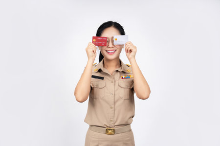 A Thai female government official in a khaki uniform holds a credit card and uses a mobile phone application against an isolated backgroundの写真素材