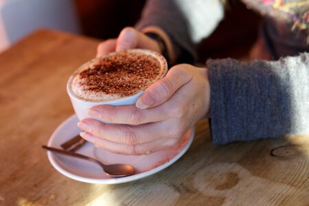 Hands holding a cup of coffee with a spoon and saucer on a wooden tableの写真素材
