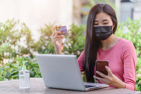 Beautiful young asian woman wearing a protective face mask hold while mobile phone paying with credit card for online shopping itemの写真素材