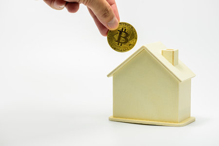 Hand places Bitcoin coins into a small wooden model of a house, symbolizing the concept of investing in real estate. using digital currency The growing trend of Bitcoin useの写真素材