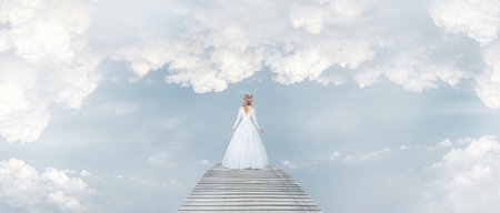 Young lady woman in white romantic wedding dress walking along fantasy bridge in cloudy skiesの写真素材