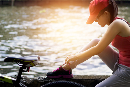 Cyclist woman sitting and tying shoeslace along the canal in sunset backgroundの写真素材