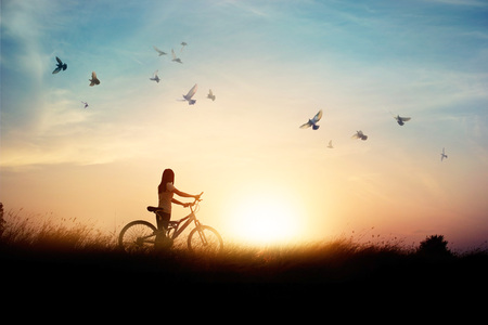 Lonely woman standing with bicycle on road of paddy field among flying birds and sunset backgroundの写真素材