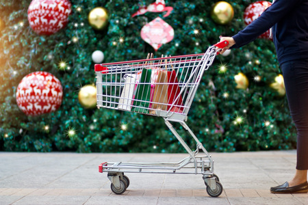 Christmas shopping - woman shopper with bags in shopping cart on holidays ornament and christmas tree on street backgroundの写真素材