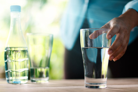 Hand touching and reaching for the glass of drinking water on wooden table, green nature background....