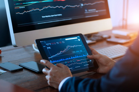 Businessman checking stock market on digital tablet and a desktop computer with stock exchange graph...
