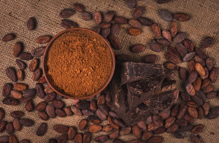 crude dark cocoa powder in a brown ceramic bowl, raw cocoa beans in the peel and raw chocolate on sacking close up, top view, ingredients for preparing chocolate and sweetsの写真素材