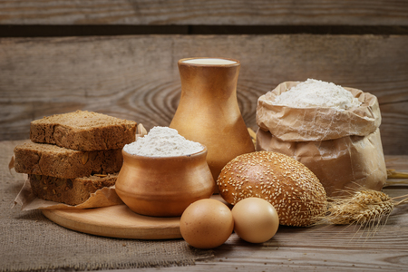 Ingredients for baking bread and pastry, milk, wheat flour, eggs, bun with sesame, rustic bread, cut into pieces, ears of wheat on the old wooden backgroundの写真素材