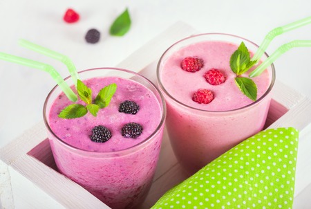 Raspberry and blackberry dairy smoothies with fresh berries and sprigs of mint, green cocktail stick in a decorative white box on a white wooden tableの写真素材