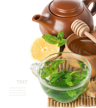 mint tea in a transparent glass cup, teapot, lemon, honey and honey spoon on a bamboo mat on a white backgroundの写真素材
