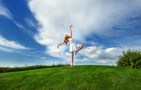 Caucasian white woman dancing in white short dress on a background of green grass and blue sky with cloudsの写真素材