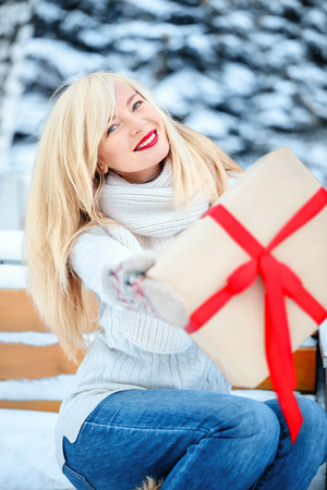 Young beautiful blond Caucasian woman 30 years in a sweater, hat and scarf with a Christmas gift in a winter park near a snow-covered treeの写真素材