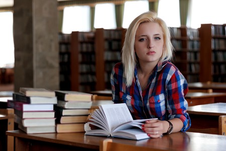 Young blonde woman sitting at the table. In the background are the rows of bookshelves. She holds in...