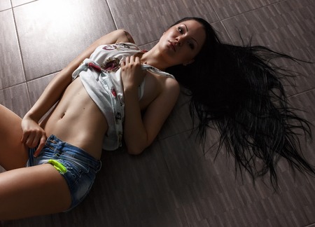 Girl with long dark hair in blue jeans shorts lying on the floor. She shows her sexy body and bellyの写真素材