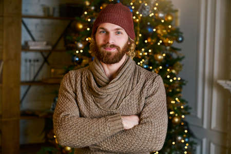 Handsome young man stands amid the Christmas tree folding hands and smiles. Fashionable man in a cozy new year apartment. Young guy in fancy sweater waiting his gift or syrprise on party.の写真素材