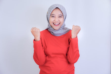 A woman wearing a grey hijab and red shirt is smiling joyfully, raising both fists in excitement.の写真素材
