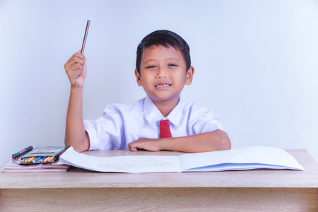 A Indonesian young boy in a school uniform, sitting at a desk with a raised hand, smiling, with books and crayons, ready to learn.の写真素材