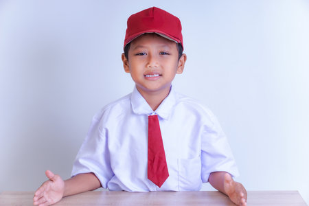 An Indonesian boy in a school uniform is smiling, sitting at a desk with both hands resting on it. He is wearing a white shirt, a red tie, and a red cap. A plain, bright background highlights him.の写真素材