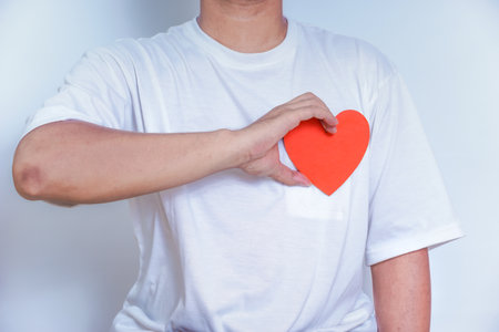 A person wearing a white shirt holds a red paper heart close to their chest, symbolizing love and...