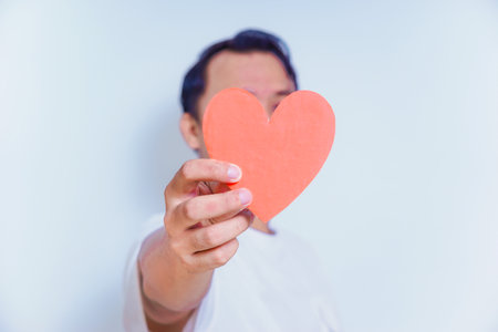 A person holds up a red heart-shaped cutout, symbolizing love and affection against a clean white background. World Heart Day conceptの写真素材