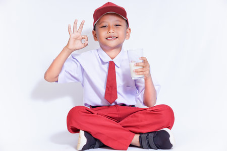 A cheerful boy in a cap and tie, sitting cross-legged, holding a glass of milk and flashing an "okay" sign.の写真素材