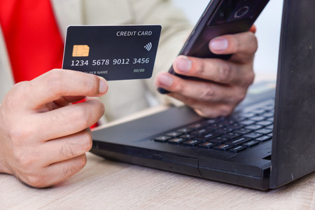 A person with red shirt holding a credit card in one hand and a smartphone in the other while near a laptop.の写真素材