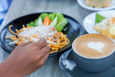 a portrait of a hand holding chopsticks to pick up pasta on a plate and a latte with a heart-shaped toppingの写真素材