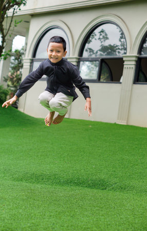 A joyful asian boy leaps in the air, showcasing playfulness and energy on a vibrant green lawn.