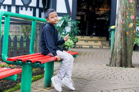 A young asian boy sits on a colorful bench, smiling and enjoying his surroundings in a park.の写真素材