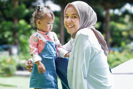A joyful asian muslim woman and her child share a moment outdoors in a vibrant garden.