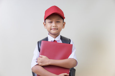 A cheerful asian boy in a red cap and uniform holding a red school reportの写真素材