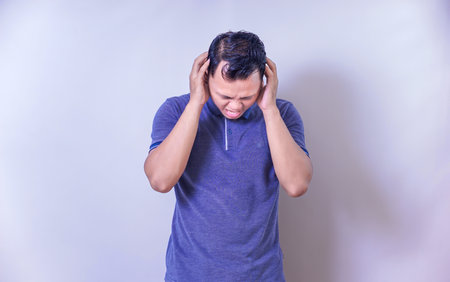 An asian man grimaces in discomfort, covering his ears to block out loud noise.