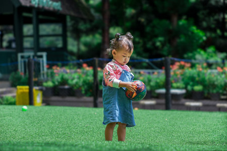 A young asian girl stands on green grass, holding a colorful ball, lost in thought amid flowers.の写真素材