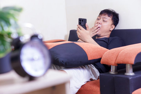 A man on a couch, yawning while scrolling through his phone, with a clock in the foreground.の写真素材