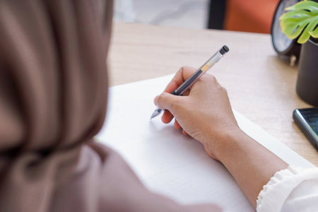 A woman in a hijab writing on paper, focusing intently at a well organized workspace.の写真素材