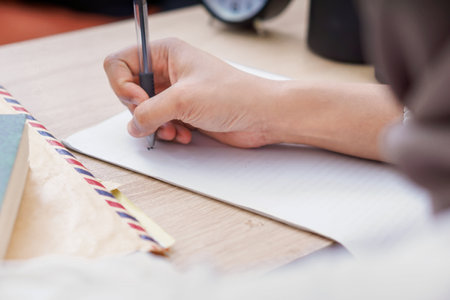 A woman hand holding a pen, writing on paper with books and a clock nearby.