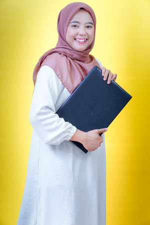 A smiling asian woman in a hijab holds a laptop against a bright yellow background, radiating confidence.の写真素材