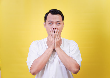 A surprised asian man covering his mouth against a bright yellow backgroundの写真素材