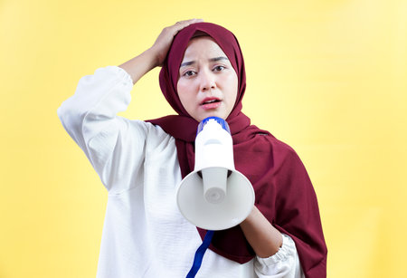 Concerned asian woman in a hijab while holding a megaphone against a yellow background.