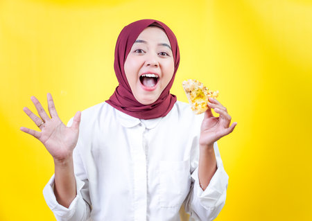 A joyful asian woman in a white outfit and hijab holds a snack against a yellow backgroundの写真素材