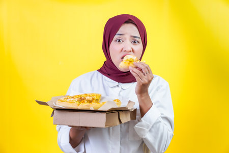 An asian woman enjoys a slice of pizza while holding a box of pizza in front of a bright yellow background.の写真素材
