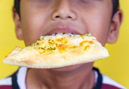 Close up of cheerful boy with a slice of pizza in his mouth, set against a vibrant yellow background. international foods day conceptの写真素材