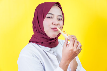 A beautiful asian woman in a hijab enjoys a slice of pizza against a bright yellow backgroundの写真素材