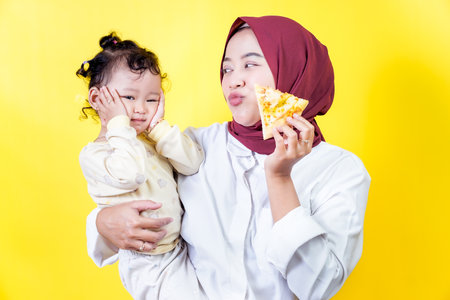 A joyful woman holds her smiling toddler and a piece of pizza against a bright yellow backgroundの写真素材