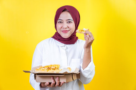 An asian joyful woman in a hijab enjoys pizza against a vibrant yellow backgroundの写真素材