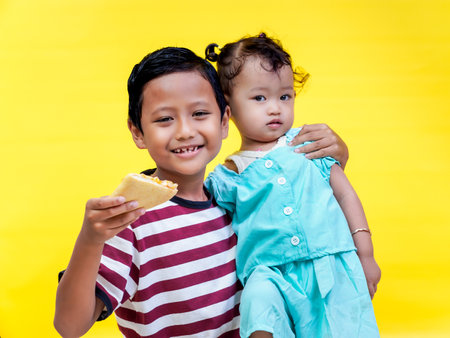 Happy siblings enjoying food together against a bright yellow background.の写真素材